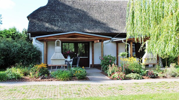 A rustic house with a thatched roof, a covered porch, and a garden with various plants and flowers. - Zur Alten Boddenfischerei (Saal)