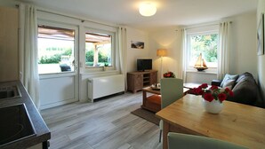 A modern kitchen with a built-in stove, a dining area with a wooden table and chairs, a TV on a wooden cabinet, and large windows with curtains. - Zur Alten Boddenfischerei (Saal)