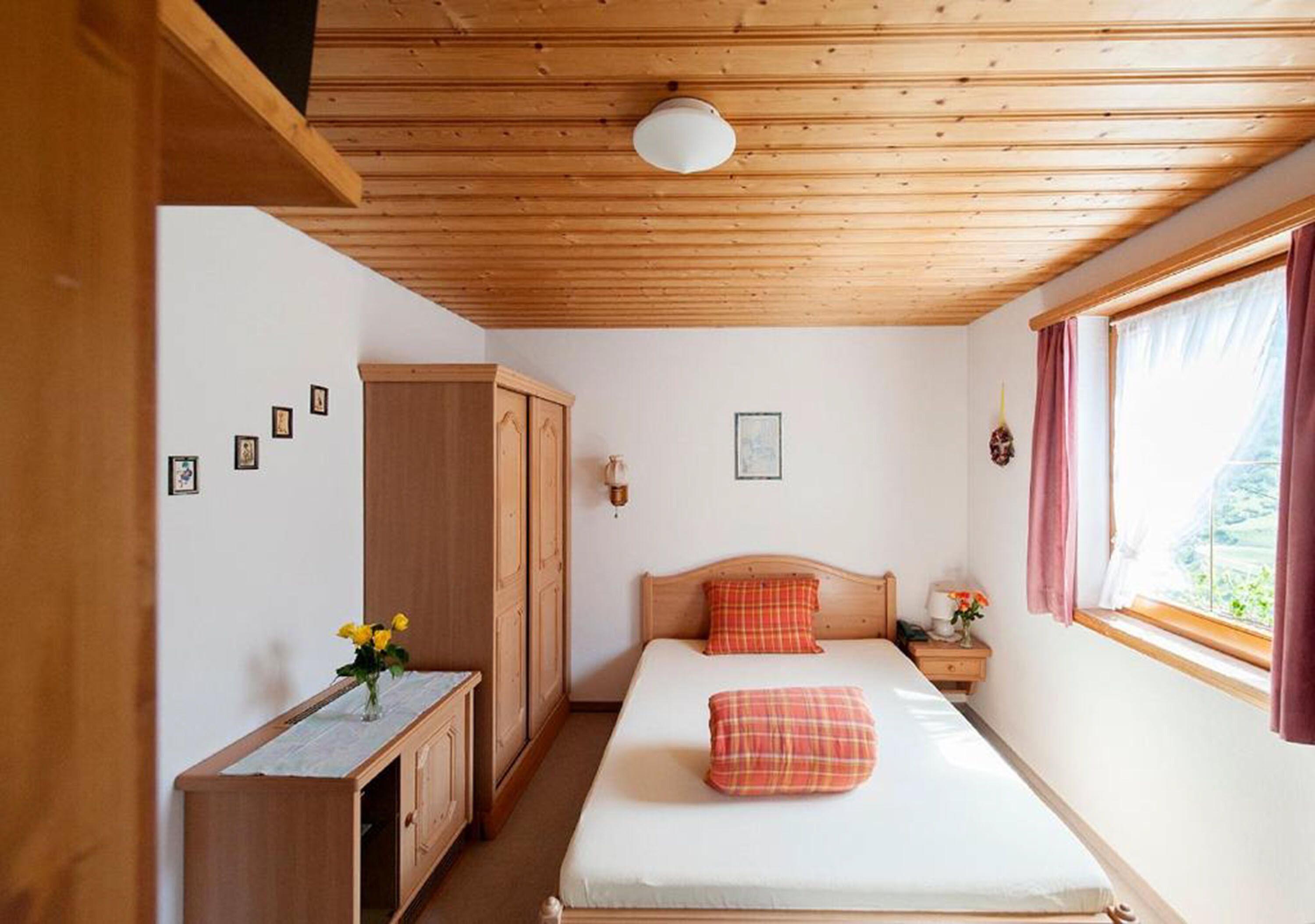 Une chambre avec un plafond en bois, un lit avec un oreiller rouge, une table de chevet, une armoire et une fenêtre avec des rideaux.