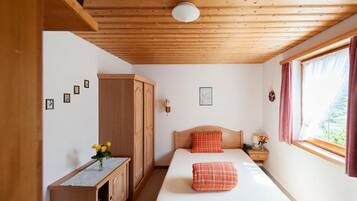 A bedroom with a wooden ceiling, a bed with a red pillow, a nightstand, a wardrobe, and a window with curtains.