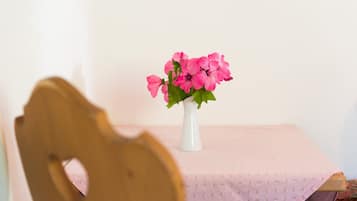 A table with a white vase containing pink flowers, a wooden chair in the foreground, and a pink tablecloth.