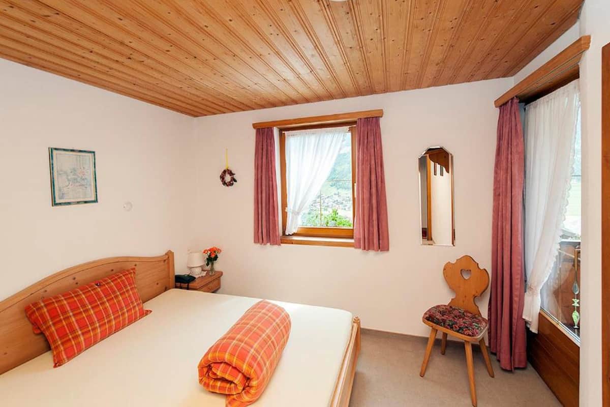 A bedroom with a wooden ceiling, a bed with a red and orange blanket, a chair with a floral cushion, a window with pink curtains, and a mirror on the wall.