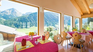 A dining area with tables set for two, large windows offering mountain views, and wooden beams on the ceiling. - Hotel Cuntera (Medel)