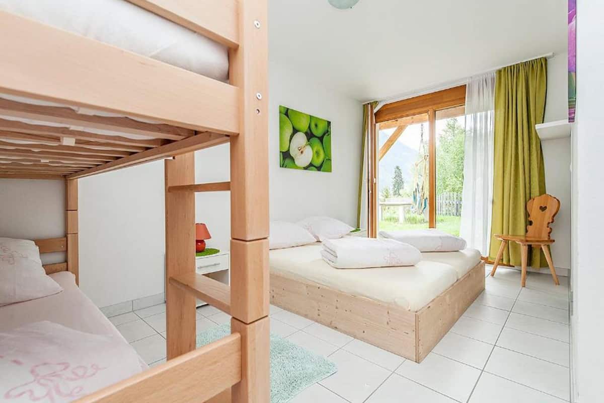 A room with a bunk bed, a single bed, a window with curtains, a painting, and a small wooden chair.