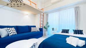 Ein modernes Hotelzimmer mit einem blauen Sofa, einem weißen runden Tisch, einem großen Bett mit blauer Bettdecke und einem Fenster mit Vorhängen.