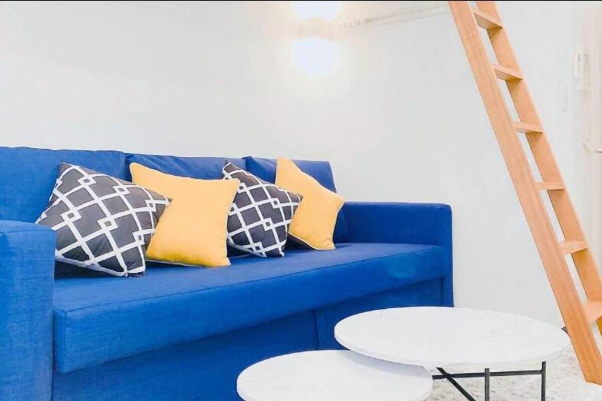 Un sofá azul con almohadones estampados, una mesa redonda blanca y una escalera de madera apoyada contra una pared blanca.