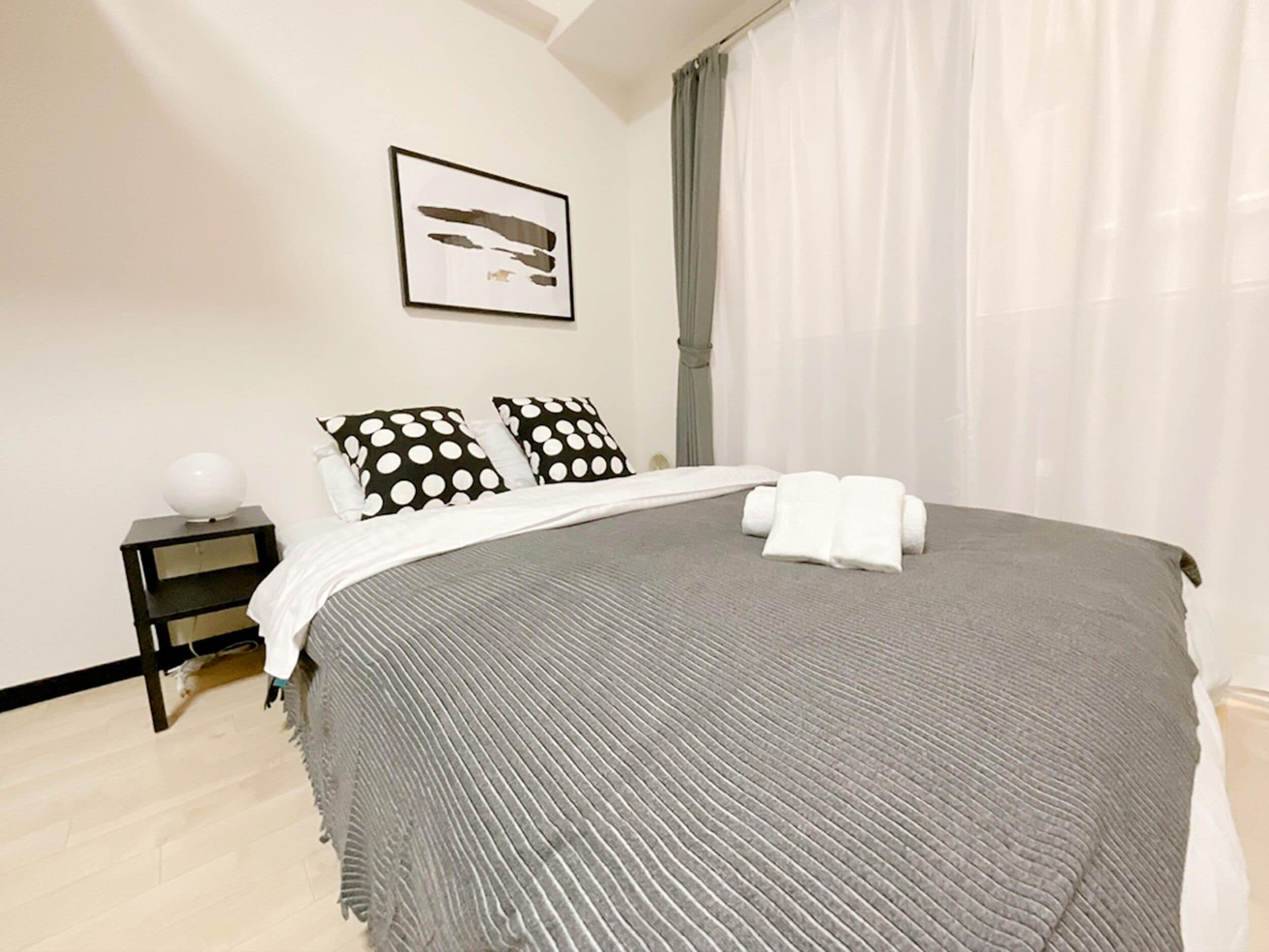 A neatly made bed with a grey blanket, two polka-dotted pillows, and a white towel folded on the bed. There is a framed picture on the wall above the bed and a small bedside table with a lamp.