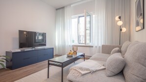 A modern living room with a sofa, a coffee table, a TV on a cabinet, and a window with curtains. - FRITZTOWER - Apartment (Mitte)