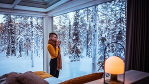 A cozy room with a large window overlooking a snowy forest, a bed with pillows, a bedside table with a candle, and a table lamp. - Arctic Skylight Lodge (Muonio)
