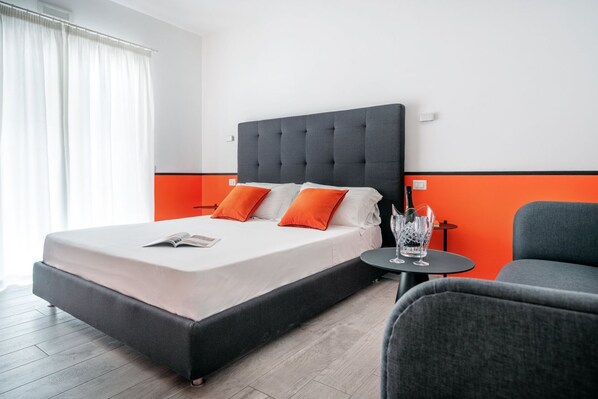 A modern hotel room with a large bed, two orange pillows, a grey upholstered armchair, and a small round table with a glass.