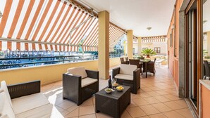 A balcony with wicker furniture, a table with a bottle and fruit, and a view of a pool and buildings. - Brand new with terrace, solarium, gym&tennis court (Sorrento)