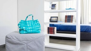 Habitació moderna amb un otomà gris, una bossa quadrada blauverd, una estant blanca amb llibres i una fotografia emmarcada, i un llit amb un mantell blau.