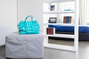 A modern room with a grey ottoman, a teal checkered bag, a white shelving unit with books and a framed picture, and a bed with a blue cover. - Brand new with terrace, solarium, gym&tennis court (Sorrento)