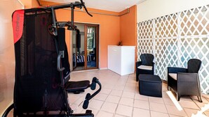 Un gimnàs petit i funcional amb equipament esportiu, un frigorífic blanc, mobles de vimet i una paret de celosies.