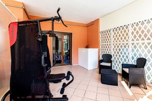 A compact home gym with exercise equipment, a white refrigerator, wicker furniture, and a lattice wall. - Brand new with terrace, solarium, gym&tennis court (Sorrento)
