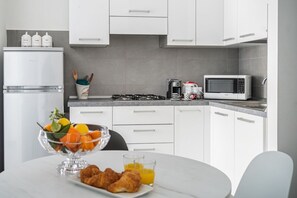 Uma cozinha moderna com armários brancos, geladeira de aço inoxidável, fogão a gás, micro-ondas e um espaço de café da manhã com croissants e suco de laranja.