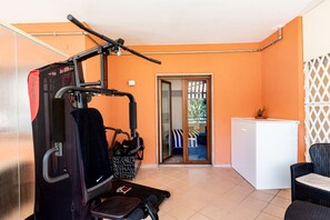 Uma academia compacta com esteira, aparelho de musculação e bicicleta ergométrica. Uma porta de vidro deslizante dá acesso a uma varanda com mobiliário de exterior.