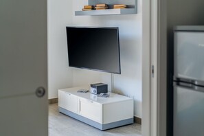 Un salon moderne avec un téléviseur à écran plat fixé au mur, un meuble blanc avec des tiroirs et une petite étagère au-dessus du téléviseur où sont rangés des livres.