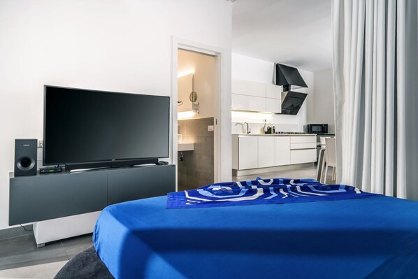 A modern hotel room with a large flat-screen TV, a bed with a blue bedspread, a kitchenette, and a bathroom visible through an open door.