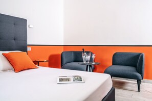 Hotellihuone, jossa on sänky, kaksi nojatuolia, pieni pöytä, jonka päällä on juoma, ja sängyllä lehti.