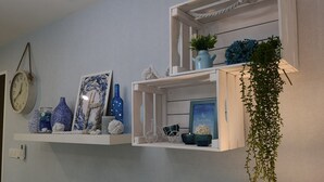 Et værelse med en lyseblå væg med struktur, et hvidt ur og en hylde med pynteting, herunder vaser, et indrammet billede og en hængende plante.