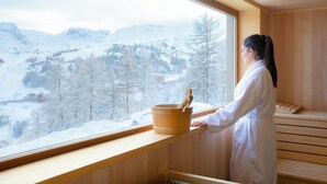 Una sauna con interior de madera, una ventana que ofrece vistas a montañas nevadas y un balde de madera con una cuchara.