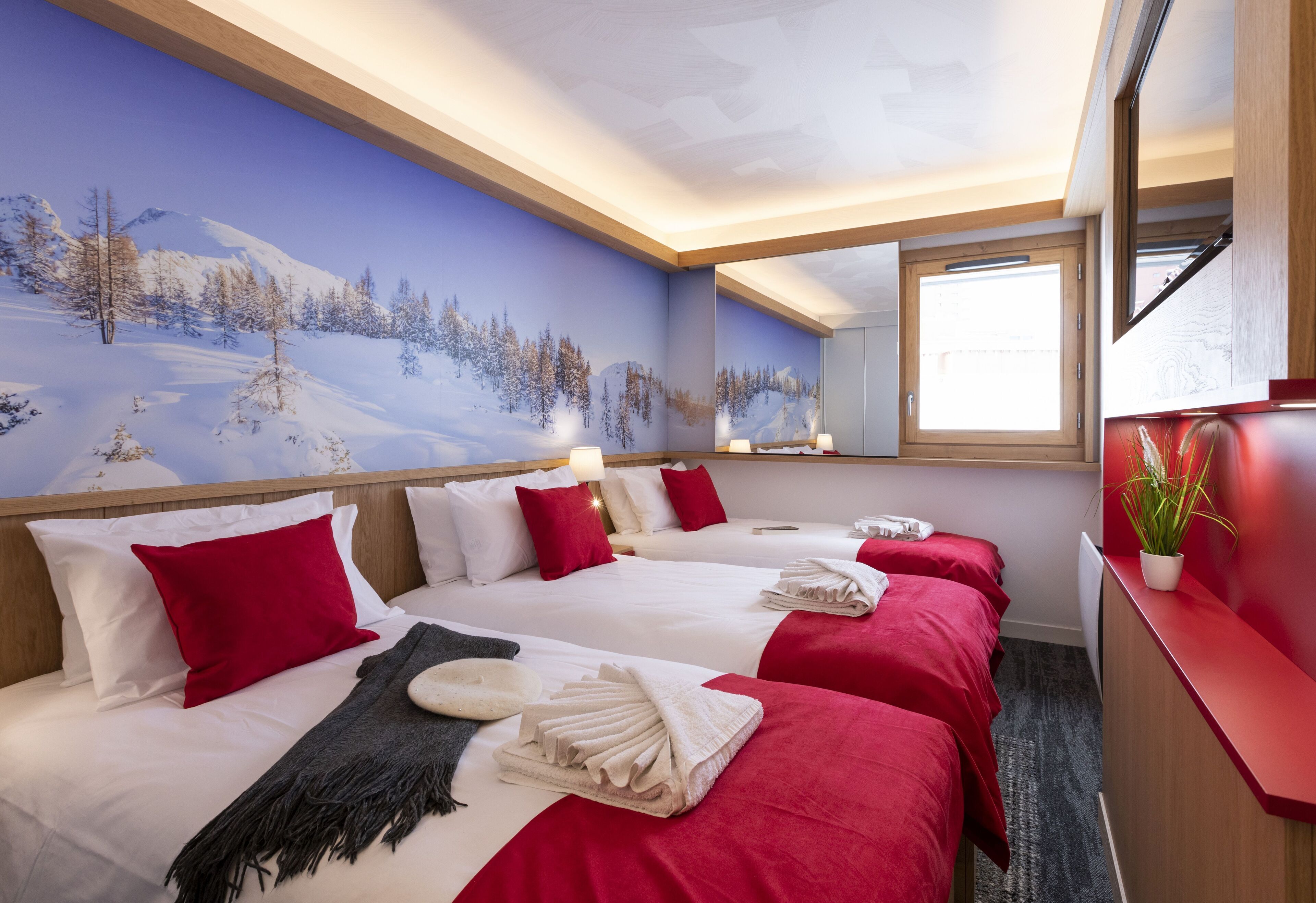 A hotel room with two beds, red and white bedding, a snowy landscape mural, a mirror, and a red desk with a plant.