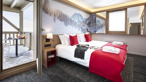 Habitación de hotel con una cama grande, vistas a montañas nevadas a través de un ventanal, una mesita con una lámpara y una manta roja.