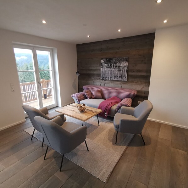 Un salón moderno con suelo de madera, sillones grises, una mesa de centro de madera y un sofá con cojines a cuadros.