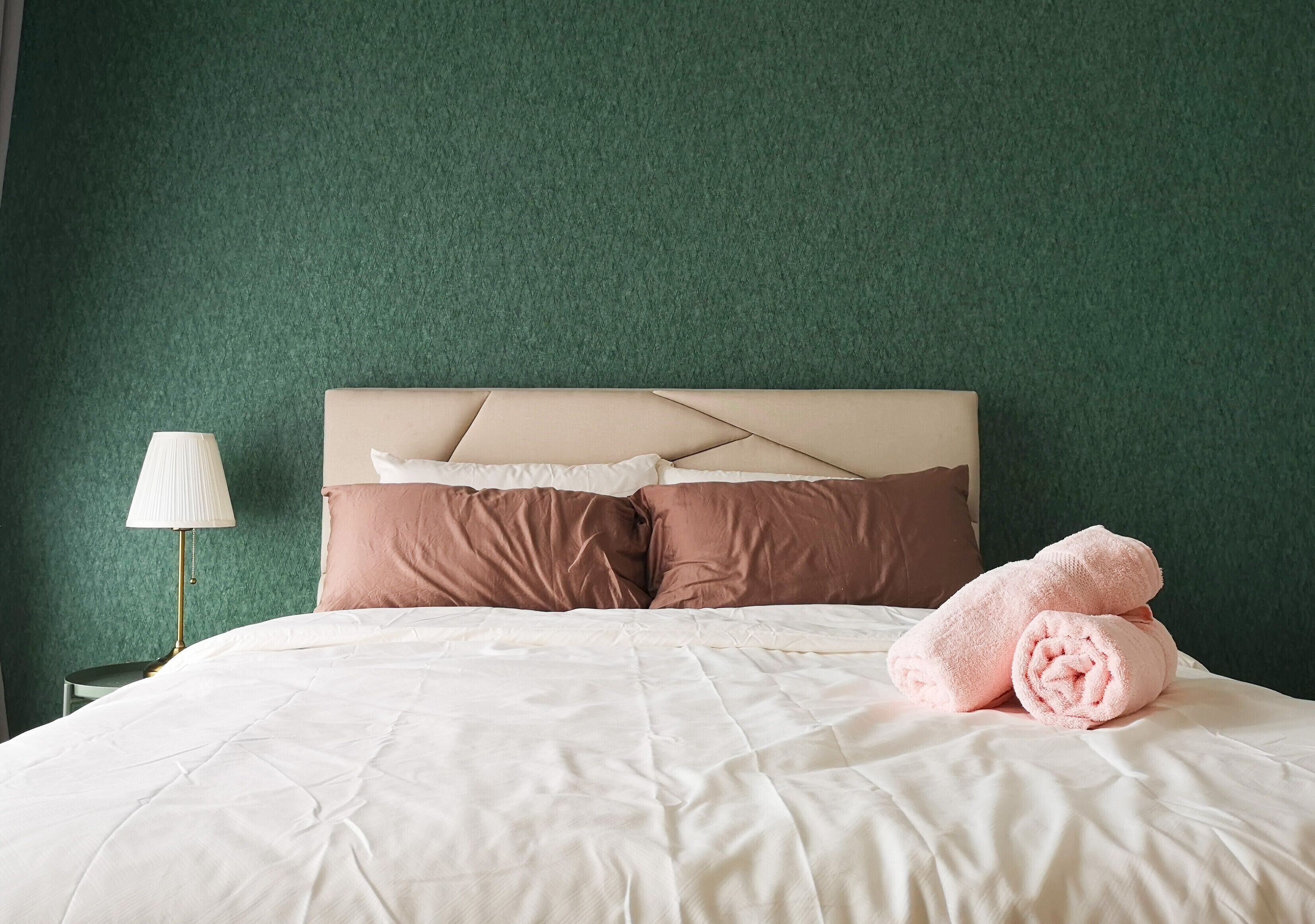 A neatly made bed with a white comforter and two brown pillows, a bedside lamp, and pink towels on the bed.