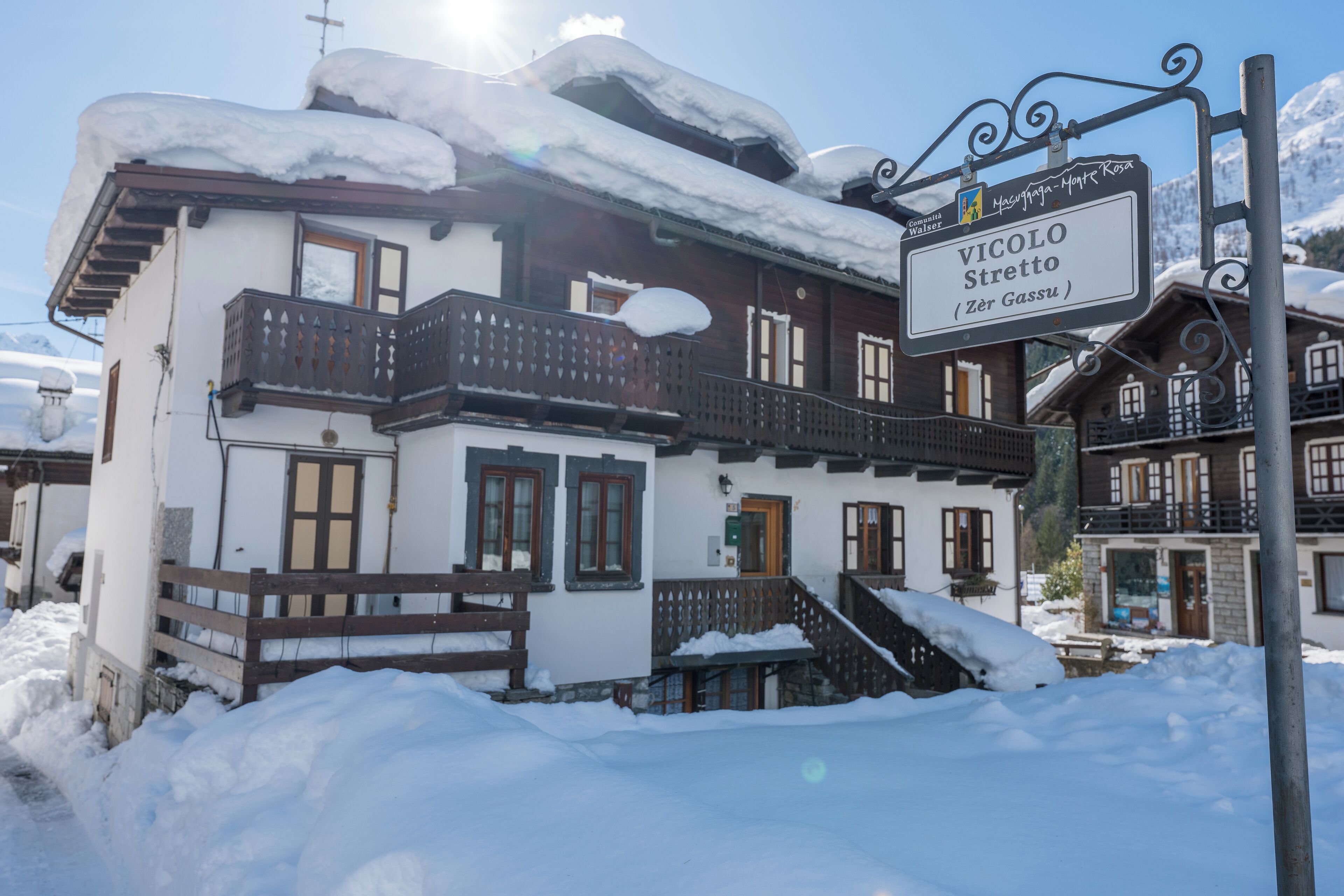 Um chalé tradicional com varanda de madeira, uma placa com o nome 'Vicolo Stretta' e uma paisagem nevada ao fundo.