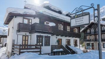 Um chalé tradicional com varanda de madeira, uma placa com o nome 'Vicolo Stretta' e uma paisagem nevada ao fundo.