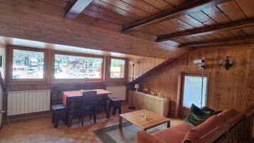 Ein gemütliches Zimmer mit Holzwänden und geneigter Decke, mit Essbereich, Tisch und Stühlen, einer Couch und einer kleinen Küchenzeile mit Arbeitsfläche und Spüle.