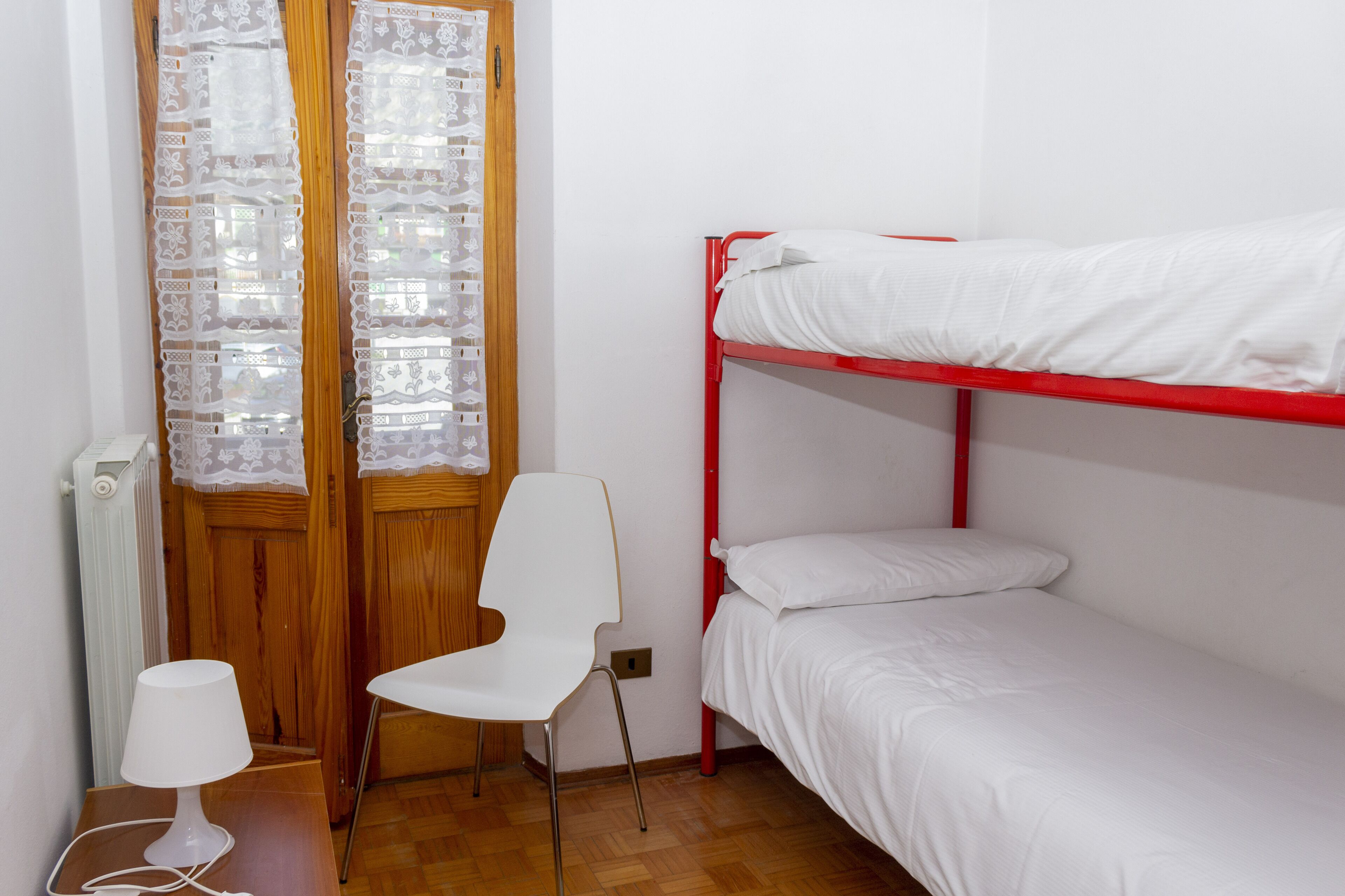 A dormitory room with a bunk bed, a chair, a desk with a lamp, and lace curtains on the door.