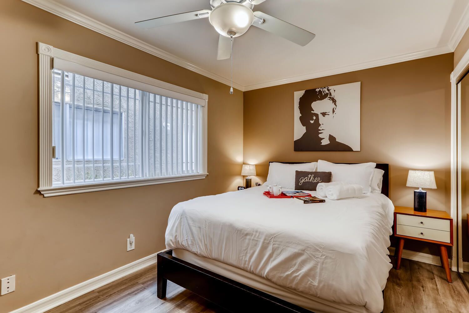 A bedroom with a large bed, a ceiling fan, a window with blinds, a bedside table with a lamp, and a wall art piece.