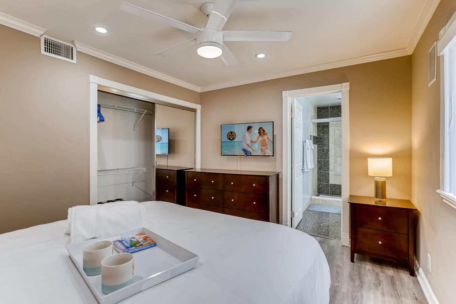 Une chambre à coucher avec un lit, une table de chevet avec une lampe, une télévision, une armoire et une salle de bain visible à travers une porte ouverte.