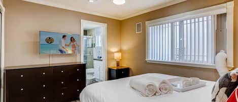 A hotel room with a bed, a TV, a dresser, a window with blinds, and a ceiling fan.