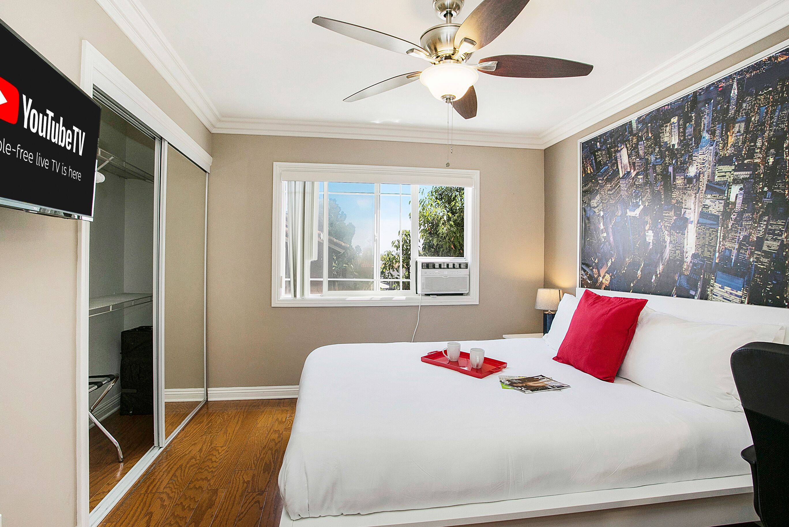 A hotel room with a bed, a ceiling fan, a window with a view of greenery, and a wall-mounted TV displaying 'YouTubeTV'.