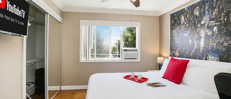 A hotel room with a bed, a ceiling fan, a window with a view of greenery, and a wall-mounted TV displaying 'YouTubeTV'.