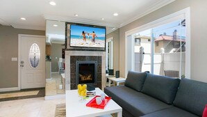 A living room with a fireplace, a large flat-screen TV, a gray sofa, and a coffee table with a tray. - Costa Mesa/Newport - Fast WiFI - Work Stations - Free Parking - Self Check In! (CM-1) (Costa Mesa)