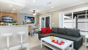 A modern kitchen with a microwave, a dining area with a table and chairs, a gray sofa with red cushions, and a white coffee table with a tray of fruit and a mug. - Awesome Multi Level - Large Patio - Fast WIFI - Work Desks - Self Check In! (CM-2) (Costa Mesa)