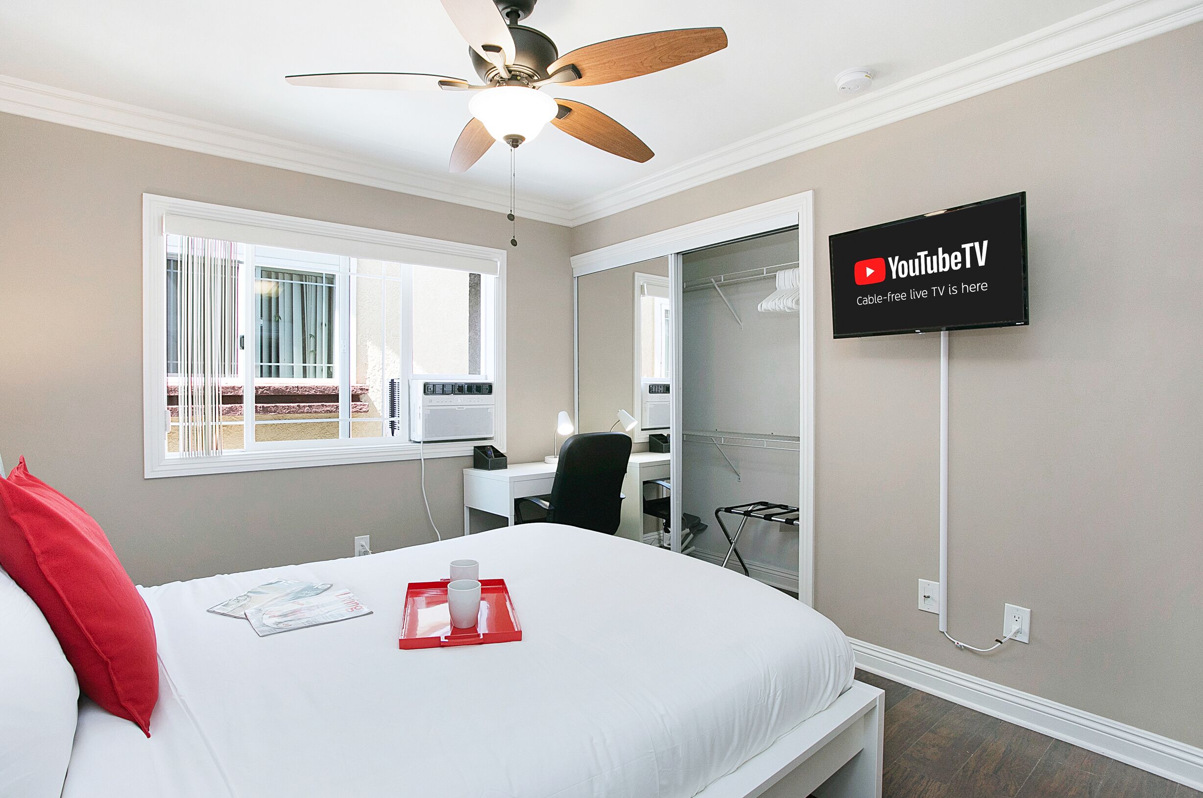 Habitación de hotel con cama, escritorio, silla, ventilador de techo, televisor montado en la pared y ventana con vista al exterior.