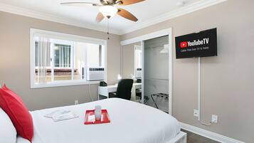 Habitación de hotel con cama, escritorio, silla, ventilador de techo, televisor montado en la pared y ventana con vista al exterior.
