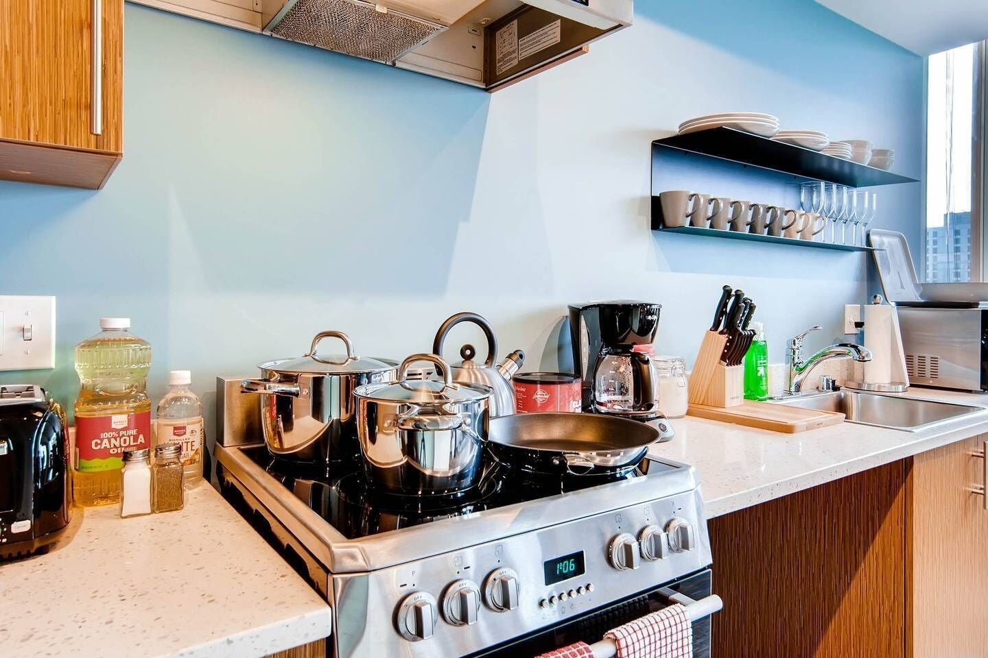 Une cuisine moderne équipée d’appareils électroménagers en acier inoxydable, notamment une cuisinière, des casseroles et une machine à café. Des étagères au-dessus du plan de travail servent à ranger la vaisselle et les verres.