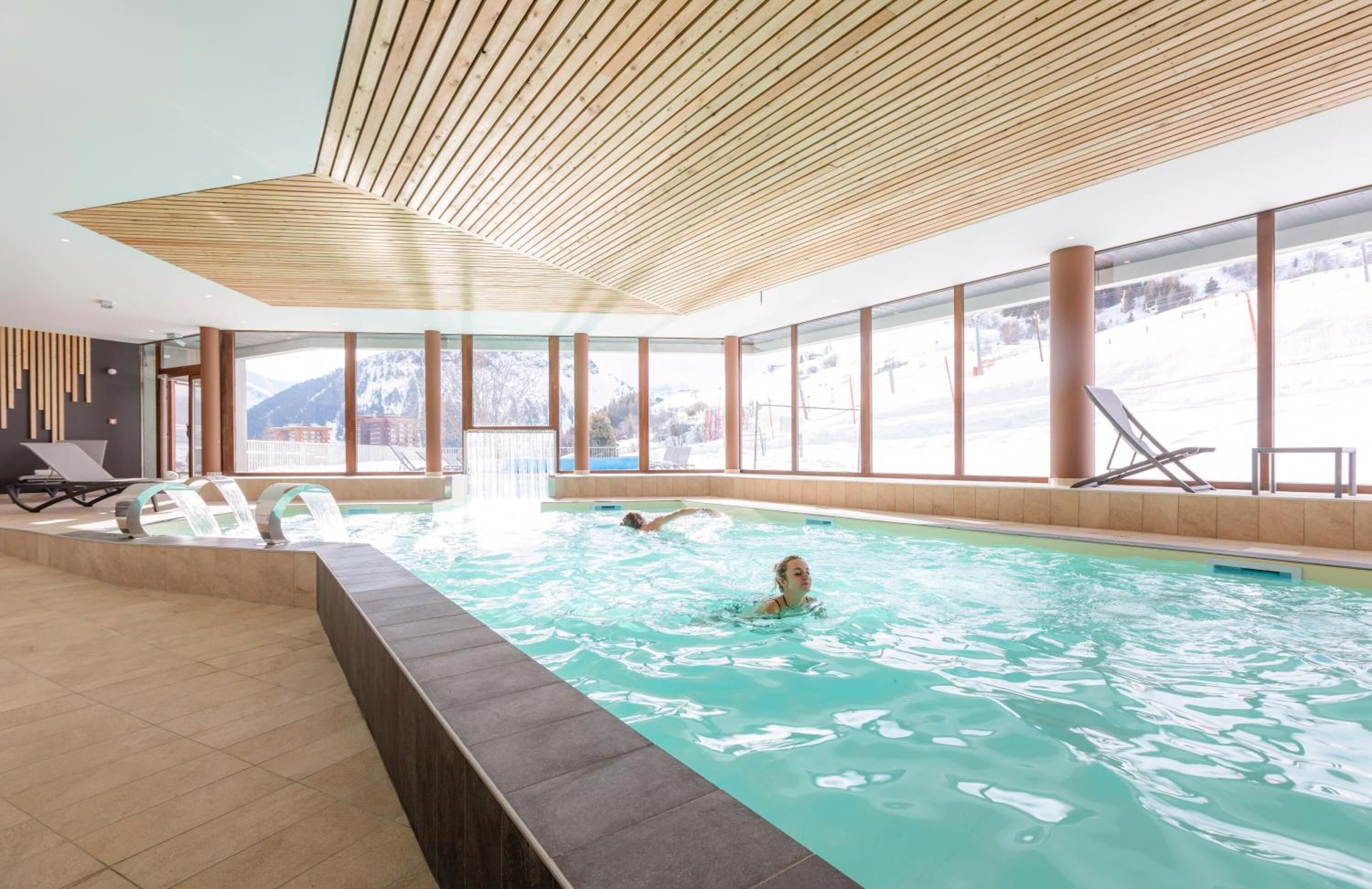 Piscina interior con vistas despejadas a las montañas, techo de madera y sillas de descanso.