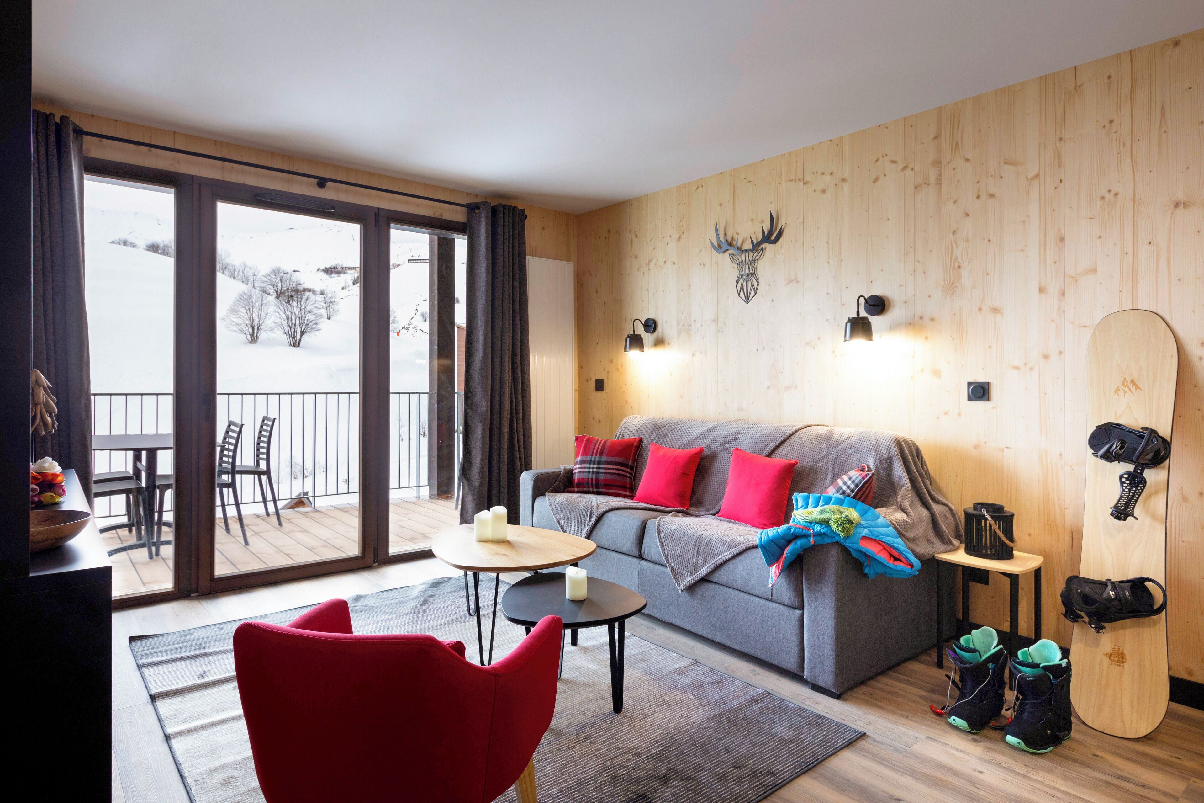 Ett mysigt vardagsrum med en soffa, en röd fåtölj och ett matbord. Ett snowboard är upphängt på väggen, och från balkongen kan man se ut över ett snötäckt landskap.