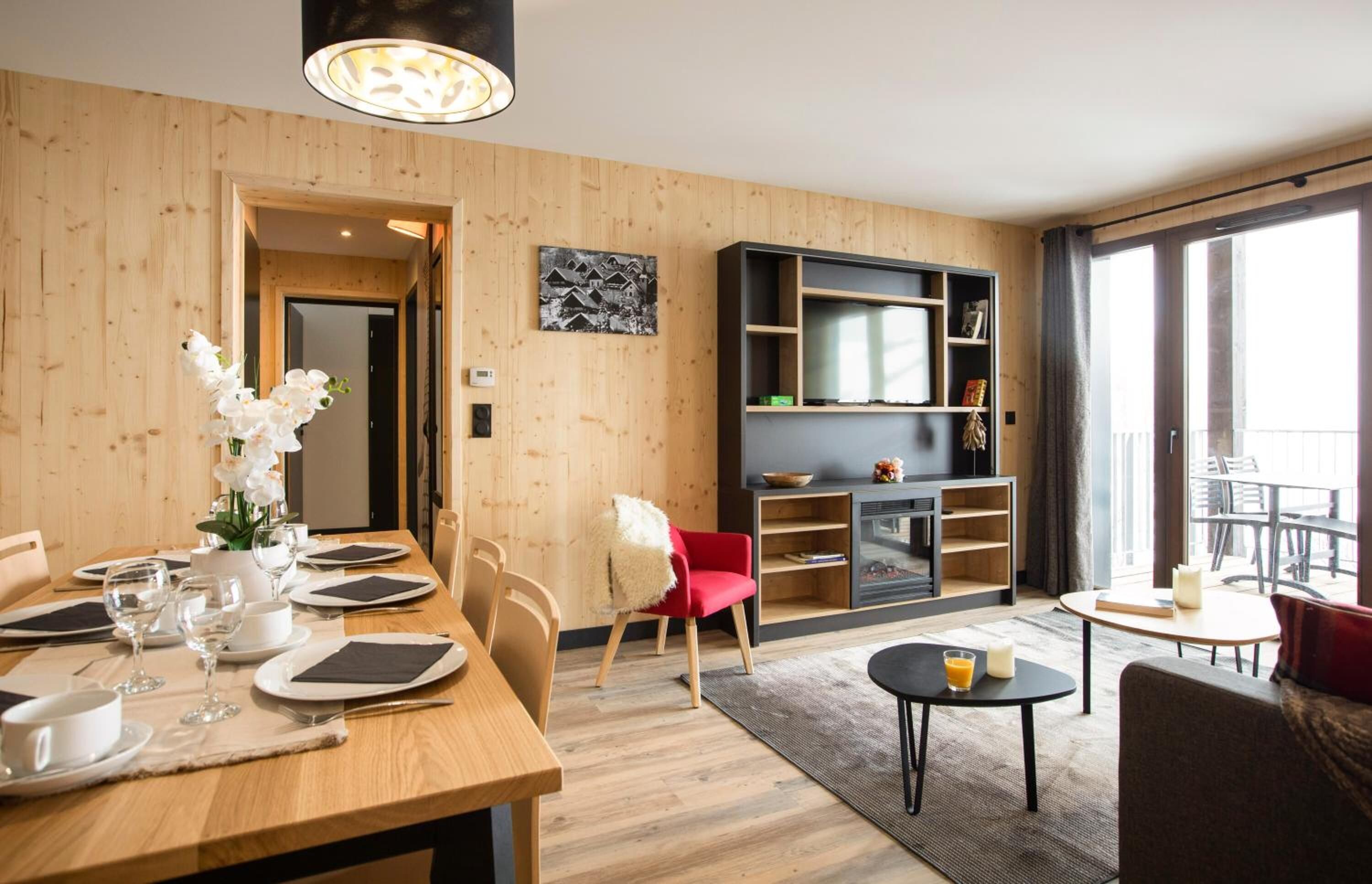 Ein modernes Wohnzimmer mit Holzboden, einem Essbereich mit Tisch für vier Personen, einem roten Sessel, einem schwarzen Sofa und einer TV-Konsole mit Regalen.