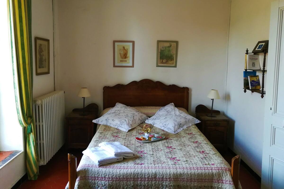 A neatly made bed with floral bedding, two pillows, and a tray with snacks. There are two wall-mounted lamps, a framed picture, and a radiator.