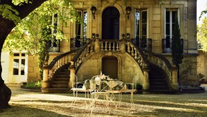 Un bâtiment historique doté d’un magnifique escalier menant à une porte, d’un espace extérieur abrité avec des tables et des chaises, et d’un grand arbre offrant de l’ombre.