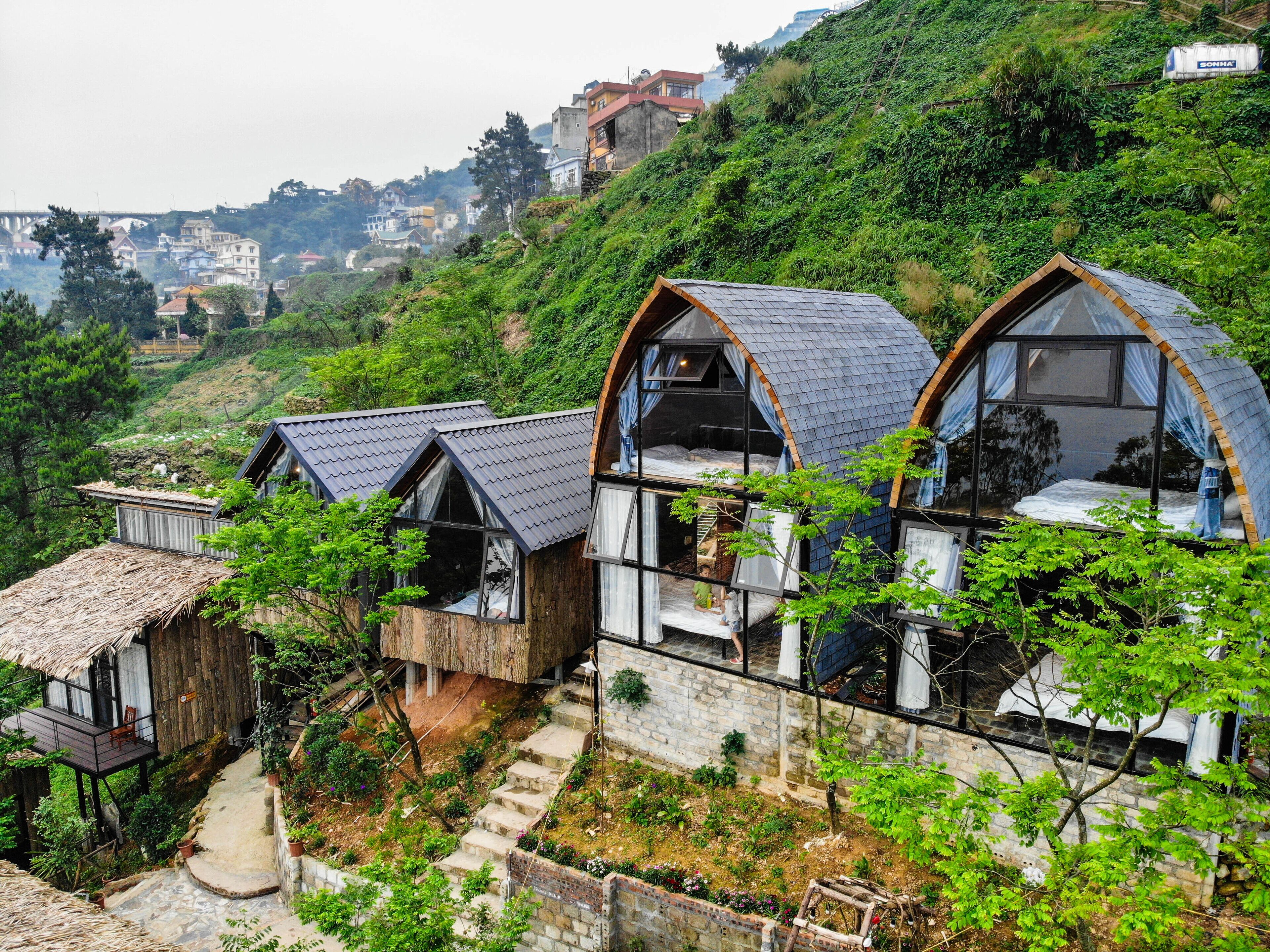 A hillside with several small, rustic cabins with steeply pitched roofs, surrounded by lush greenery and a distant view of a town.